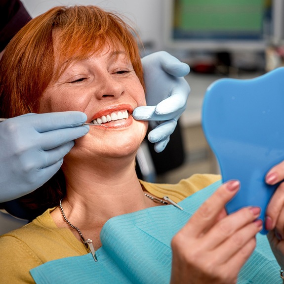 Dentist’s gloved hands touching corners of patient’s smile as she looks into mirror